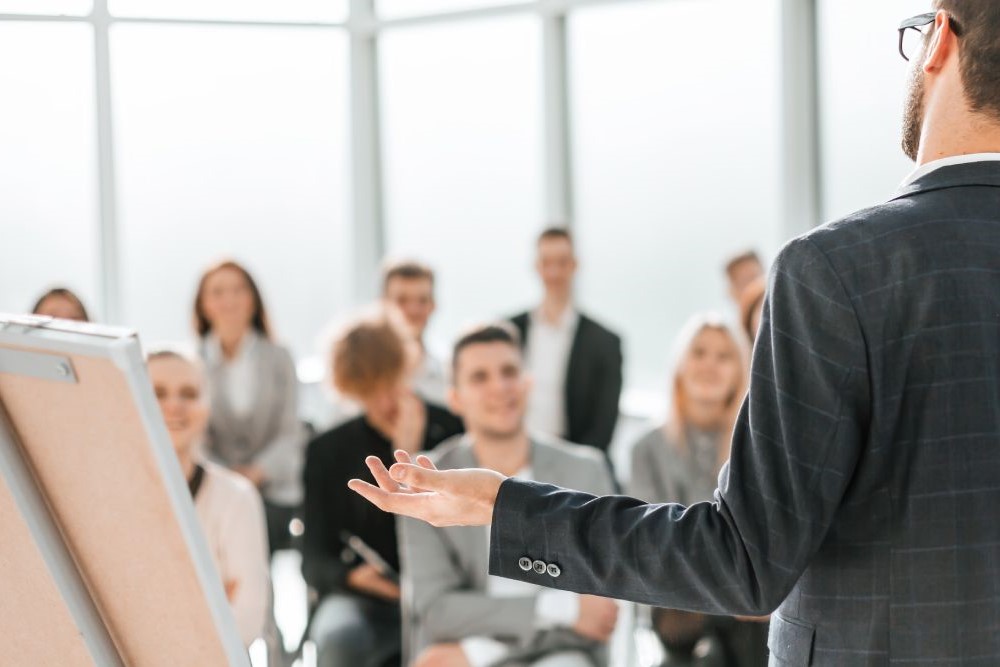 A man in a suit presenting to a group of people in a conference room.